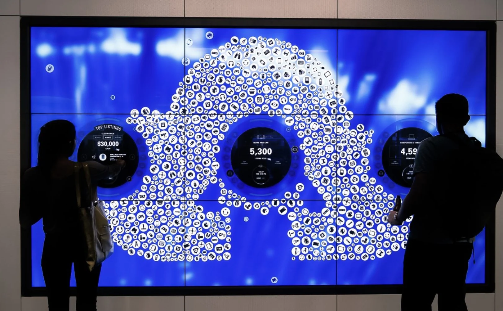 eBay Earth : deux visiteurs en silhouette devant le mur vidéo bleu, chacun avec son propre menu circulaire, autour d'un nuage de pastilles d'objets
