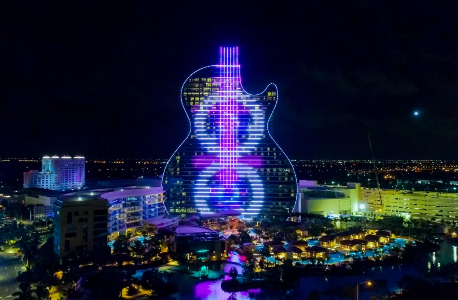 Le Guitar Hotel du Seminole Hard Rock Hotel & Casino de nuit, vu du ciel : la tour en forme de guitare illuminée de violet et de bleu au-dessus du complexe et de ses bassins