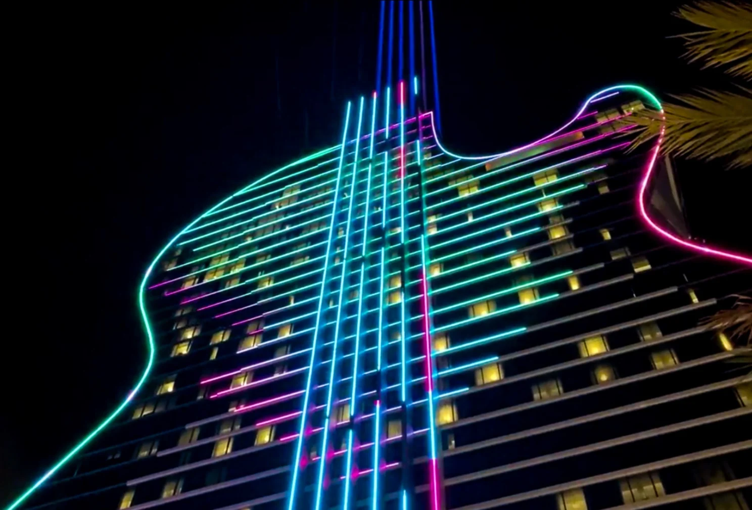 La façade du Guitar Hotel vue d'en bas, de nuit : lignes de lumière vertes, roses et bleues le long des étages, cordes bleues verticales, une palme au premier plan