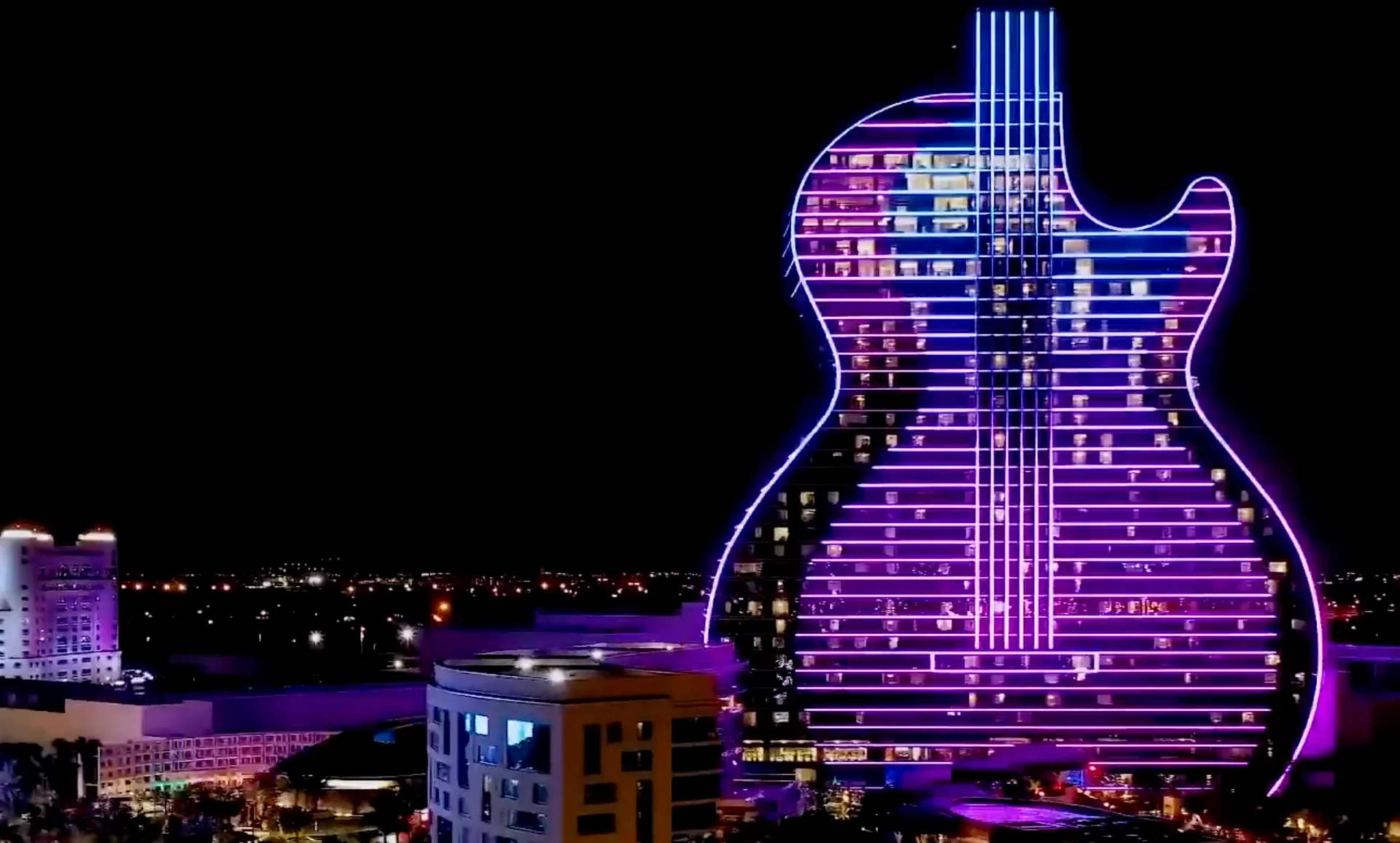 La façade du Guitar Hotel de profil, de nuit : les lignes de lumière violettes et bleues dessinent le corps et les cordes de la guitare